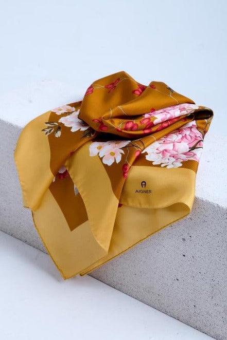 A luxury silk scarf in golden ochre, featuring a romantic pink hydrangea and white floral print, artfully bunched and draped over a white textured stone pedestal in a studio.