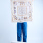 A person in blue jeans holding up a large square cream silk scarf, revealing a complete vintage-style design with ornate gold filigree borders and detailed violet illustrations of figures and landscapes against a neutral studio background.