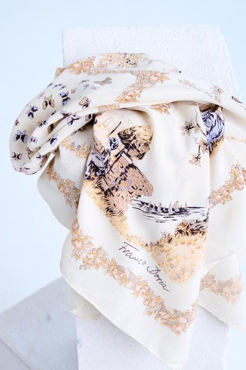A high-quality close-up of a draped silk scarf on a white stone pedestal, an intricate vintage-style print with a pastoral landscape, gold floral borders, and multi-colored butterfly motifs on a cream base.