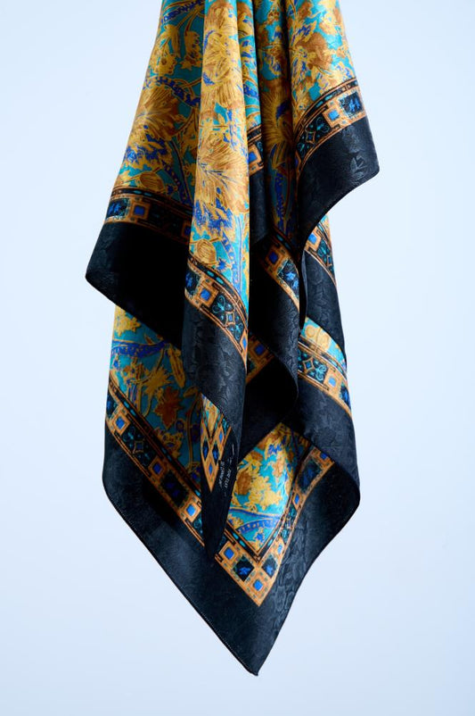 A luxury silk scarf with an ornate Baroque-inspired floral pattern in gold and turquoise blue, featuring an intricate geometric border and a bold black jacquard edge, elegantly draped in a studio setting.