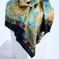A luxury silk scarf a, featuring an ornate Baroque-inspired floral pattern in gold and turquoise blue, artfully draped over a white stone pedestal to highlight its intricate geometric border and black edge.