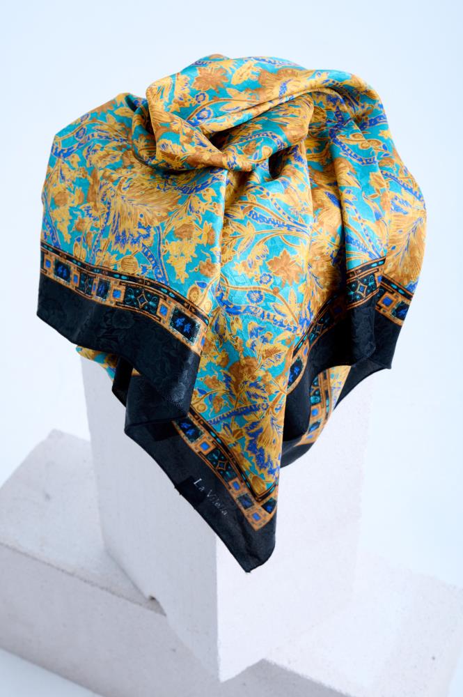 A luxury silk scarf a, featuring an ornate Baroque-inspired floral pattern in gold and turquoise blue, artfully draped over a white stone pedestal to highlight its intricate geometric border and black edge.