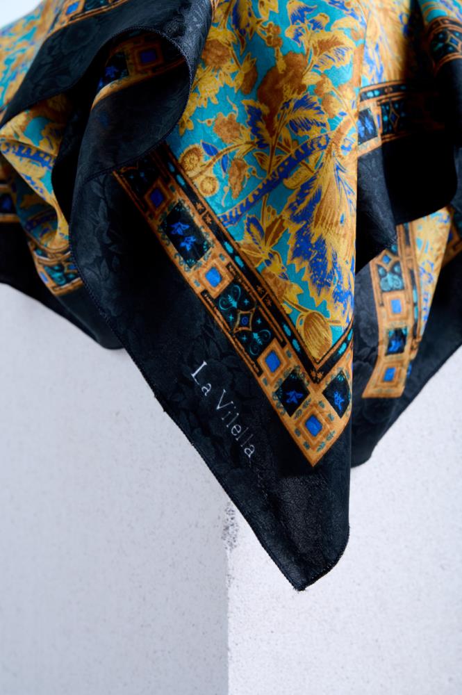 Close-up of silk scarf, showing the elegant brand signature on a black jacquard border with intricate turquoise and gold Baroque floral patterns in the background.