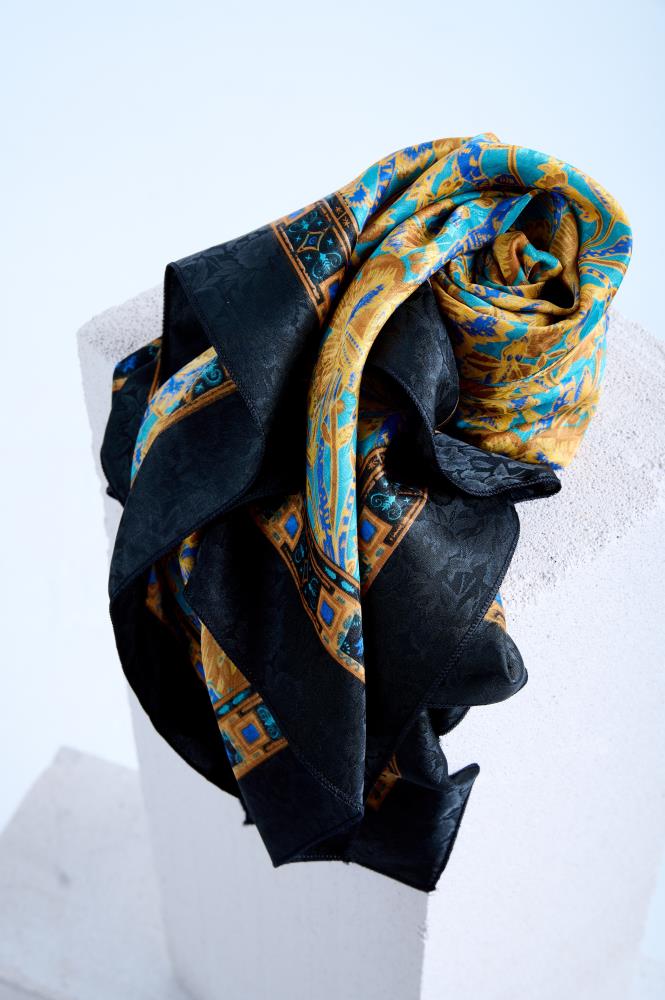 A luxury silk scarf artfully rolled and draped over a white stone pedestal, showcasing a rich turquoise and gold Baroque floral print contrasted with a wide black jacquard border featuring a subtle floral weave.