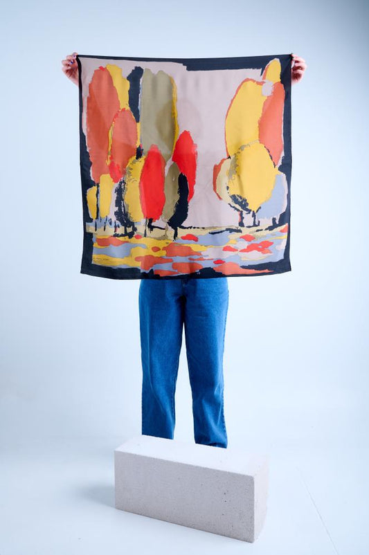 A person in blue jeans holding up a large square silk scarf featuring a vibrant Fauvist-style landscape print with stylized red, orange, and yellow trees against a neutral background, framed by a bold black border.
