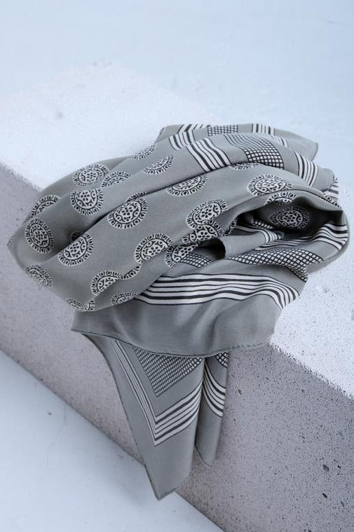 A high-angle close-up view of a high-quality grey silk scarf, featuring a classic print with repeated paisley medallions, elegant striped borders, and a fine grid motif, draped over a textured white stone edge.