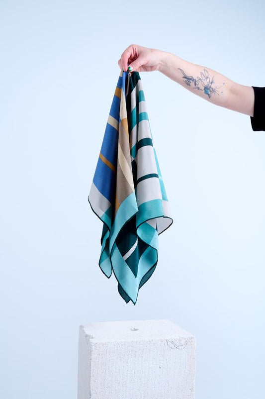 A person holding a silk scarf with a geometric pattern in shades of blue, green, and brown, featuring rolled and tucked edges.