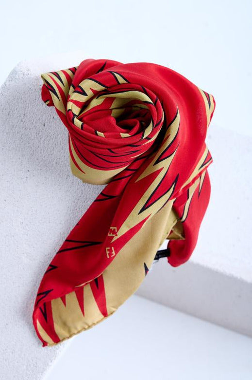 A luxury Fendi silk scarf in vibrant red and beige, artfully rolled into a rose shape to showcase its bold lightning bolt pattern and the iconic FF logo, displayed on a white textured surface.