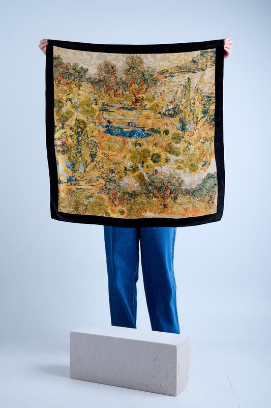 A person holding a silk scarf with black borders and a multicolored floral pattern in the center, displayed over a cement block.