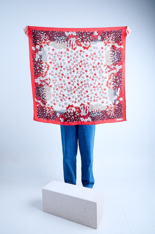 A person holding up a large square silk scarf featuring a dense floral and botanical print in red, black, and grey on a white background, finished with a solid bright red border.