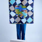 A person holding up a large square silk scarf featuring a navy blue harlequin diamond pattern in light blue and grey, with a central abstract colorful art print in green, yellow, and blue tones.