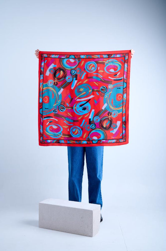 A person holding up a large square silk scarf with a vibrant abstract geometric print featuring colorful circles and strokes in red, teal, brown, and pink on a bold red background, finished with a black decorative border.