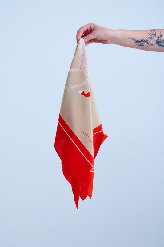 A hand holding up a beige and red silk scarf featuring a minimalist graphic design with orange accents and a bold red border, held against a neutral studio background.