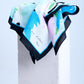 A luxury scarf with an abstract pastel print in blue, pink, and green, featuring a bold black border, elegantly draped over a white stone pedestal.