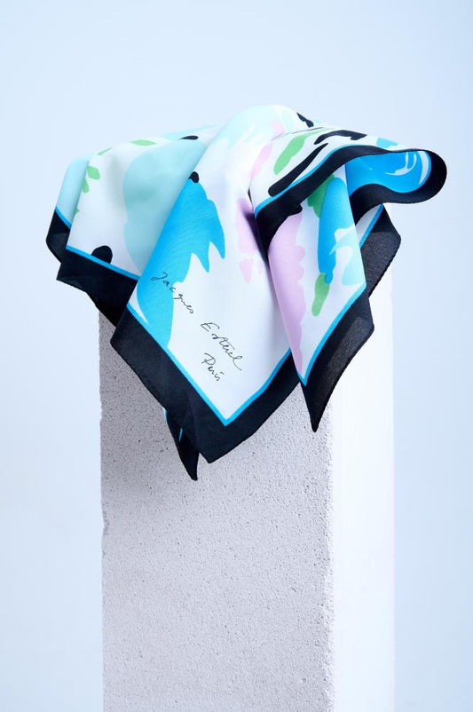 A luxury scarf with an abstract pastel print in blue, pink, and green, featuring a bold black border, elegantly draped over a white stone pedestal.