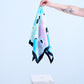 A hand with a tattoo holding up a silk scarf, featuring an abstract pastel pink and blue artistic print with a bold black border above a white stone pedestal.