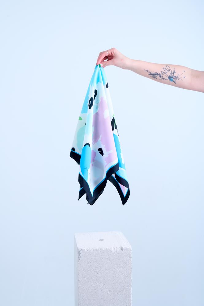 A hand with a tattoo holding up a silk scarf, featuring an abstract pastel pink and blue artistic print with a bold black border above a white stone pedestal.