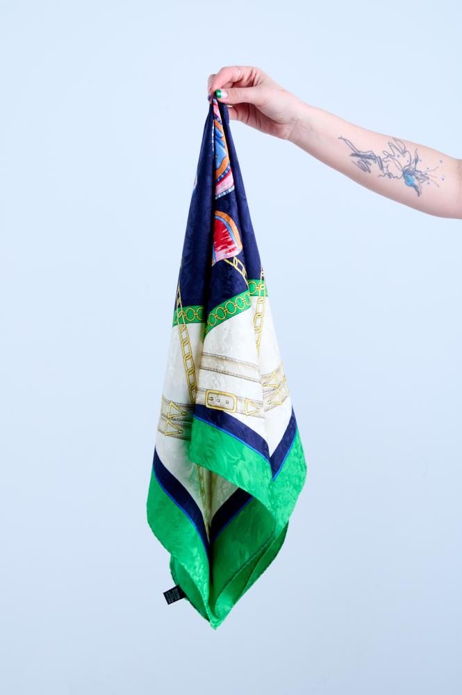 A hand holding up a luxury silk scarf with a nautical and equestrian theme, featuring golden chains, belts, and buckles on a navy blue, white, and vibrant green color-blocked background.