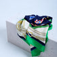 A luxury silk scarf with a classic equestrian and nautical theme, featuring golden chains and leather belt prints on a navy blue, white, and emerald green background, draped over a white stone block.