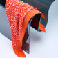 A luxury Christian Dior silk scarf in vibrant orange, featuring a colorful dot pattern in red and pink tones, elegantly draped over a modern curved glass chair.