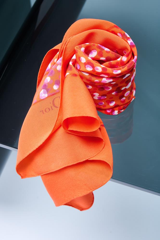 silk scarf in vibrant orange, artfully rolled to showcase a colorful mosaic-style dot pattern in pink and red tones on a reflective surface.