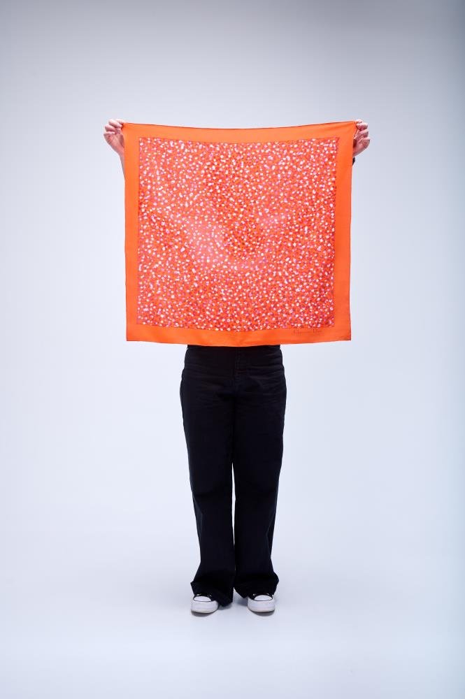 A person holding up a large square silk scarf in vibrant orange, featuring a solid border and a dense, colorful mosaic or confetti-style pattern in red, pink, and white