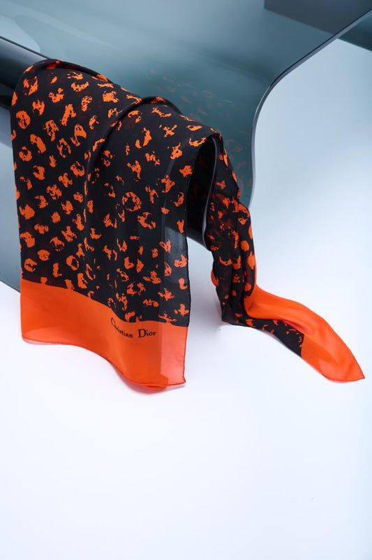 A black and orange silk scarf with a vintage Christian Dior label, displayed on a surface with a reflective background.
