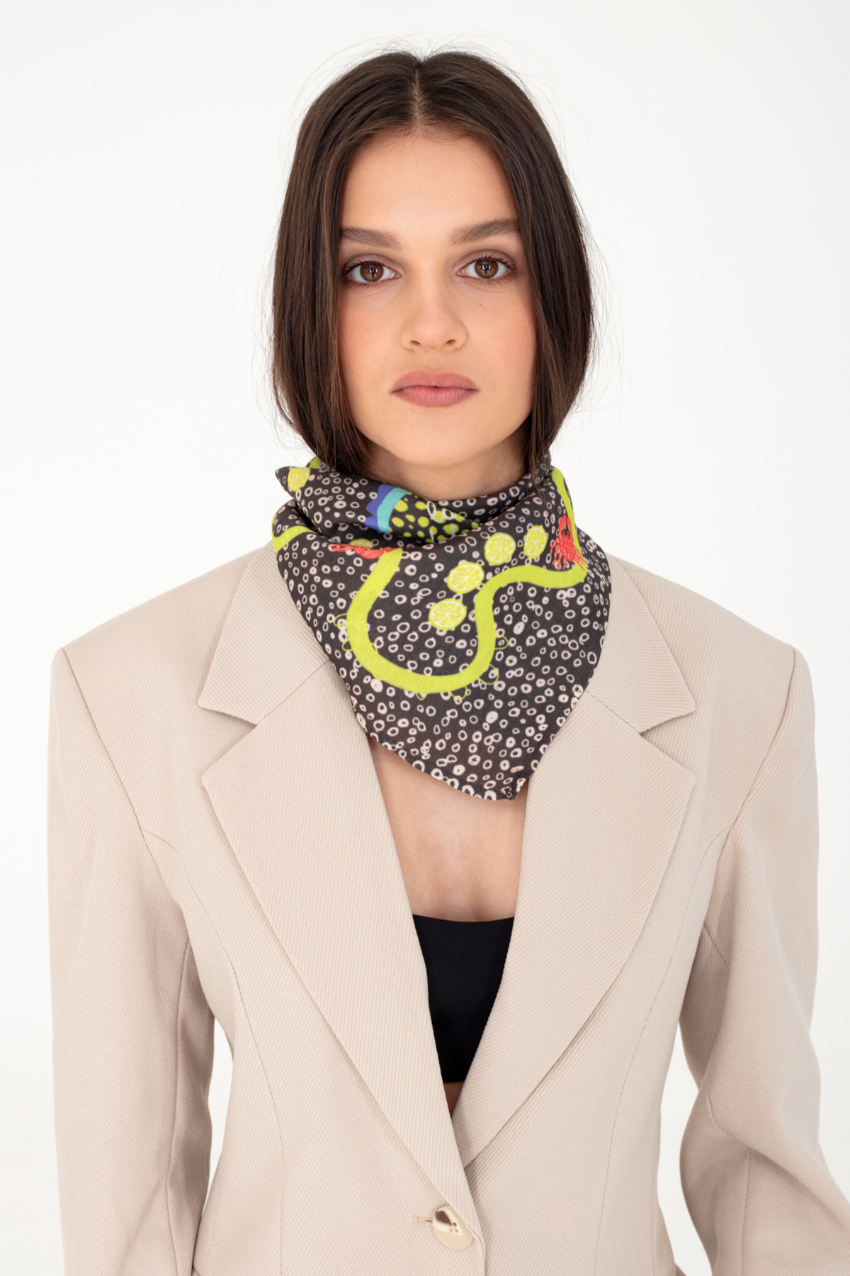 Fashion portrait of a woman wearing the "La Dolce Vita" dark brown patterned silk and viscose scarf featuring yellow lemon slice illustrations, styled with a light beige blazer against a plain white studio background