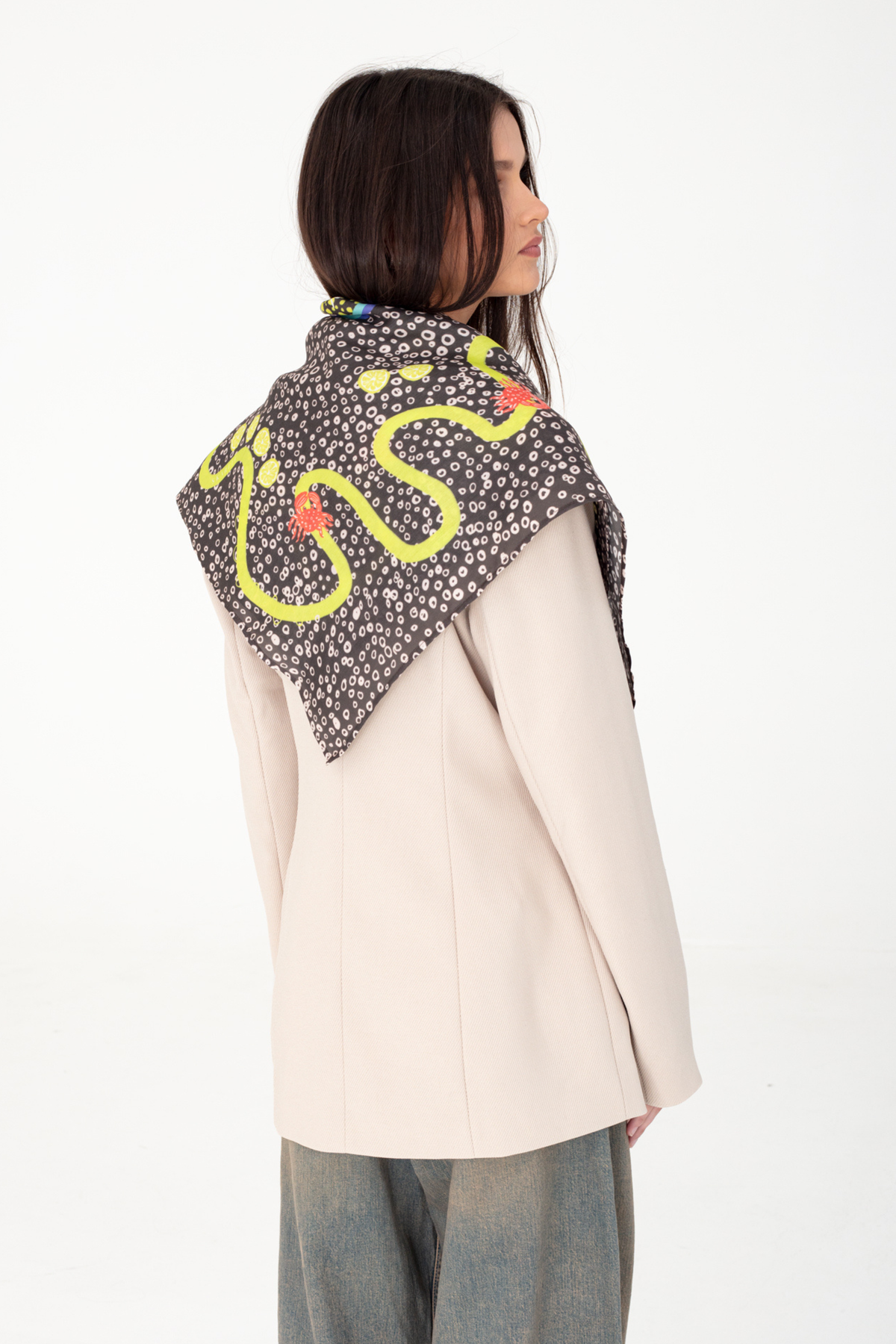 Back three-quarter perspective portrait showing a dark brown patterned silk and viscose scarf with yellow lemons and red lobsters draped triangularly over a light beige blazer, paired with blue-grey denim trousers, seen from the side-rear on a white studio background
