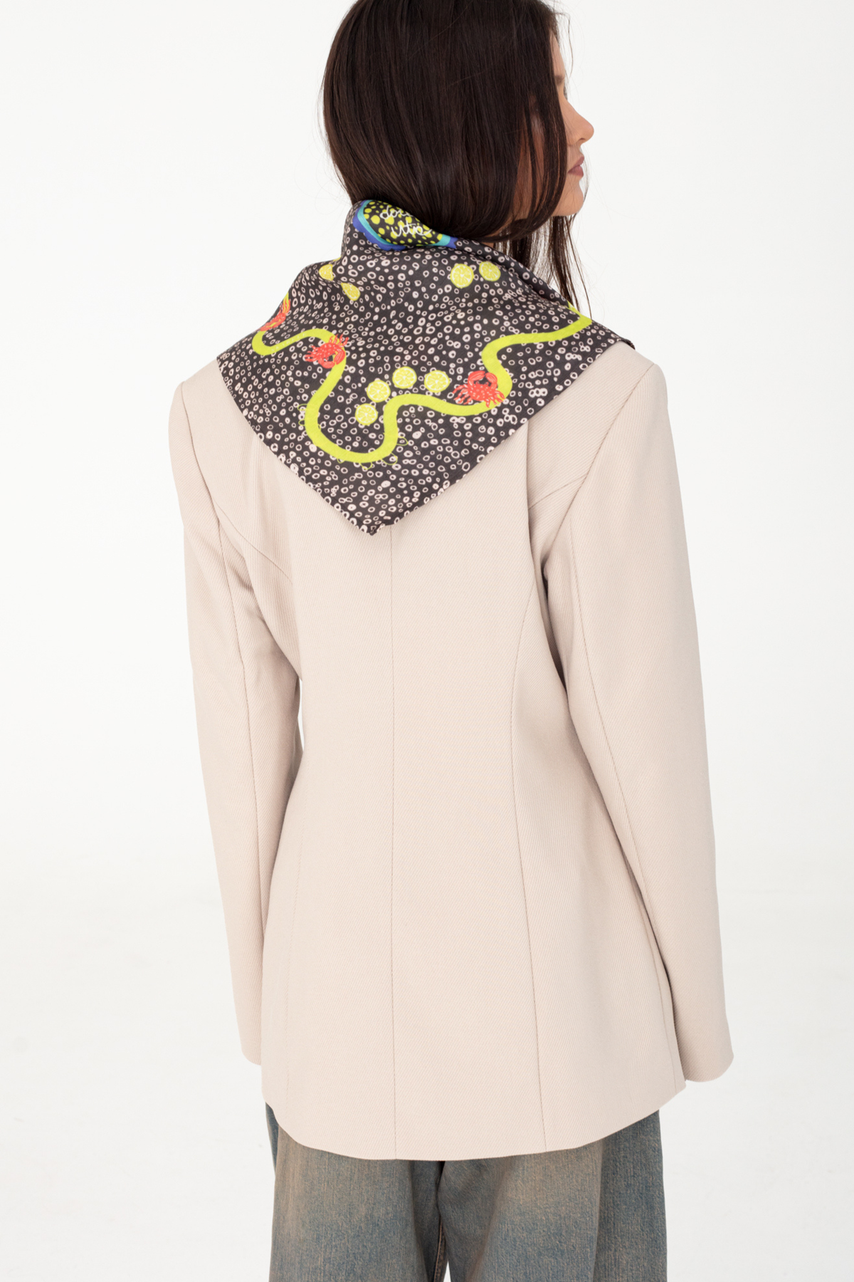 Back view fashion portrait showing a dark brown patterned silk and viscose scarf with yellow lemon and red lobster motifs draped triangularly over a light beige blazer, paired with blue jeans against a plain white studio background.