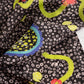 A close-up of a patterned silk and viscose fabric with a dark brown background, white circular spots, bright yellow-olive green wavy lines, red crabs, and blue and yellow circle motifs.