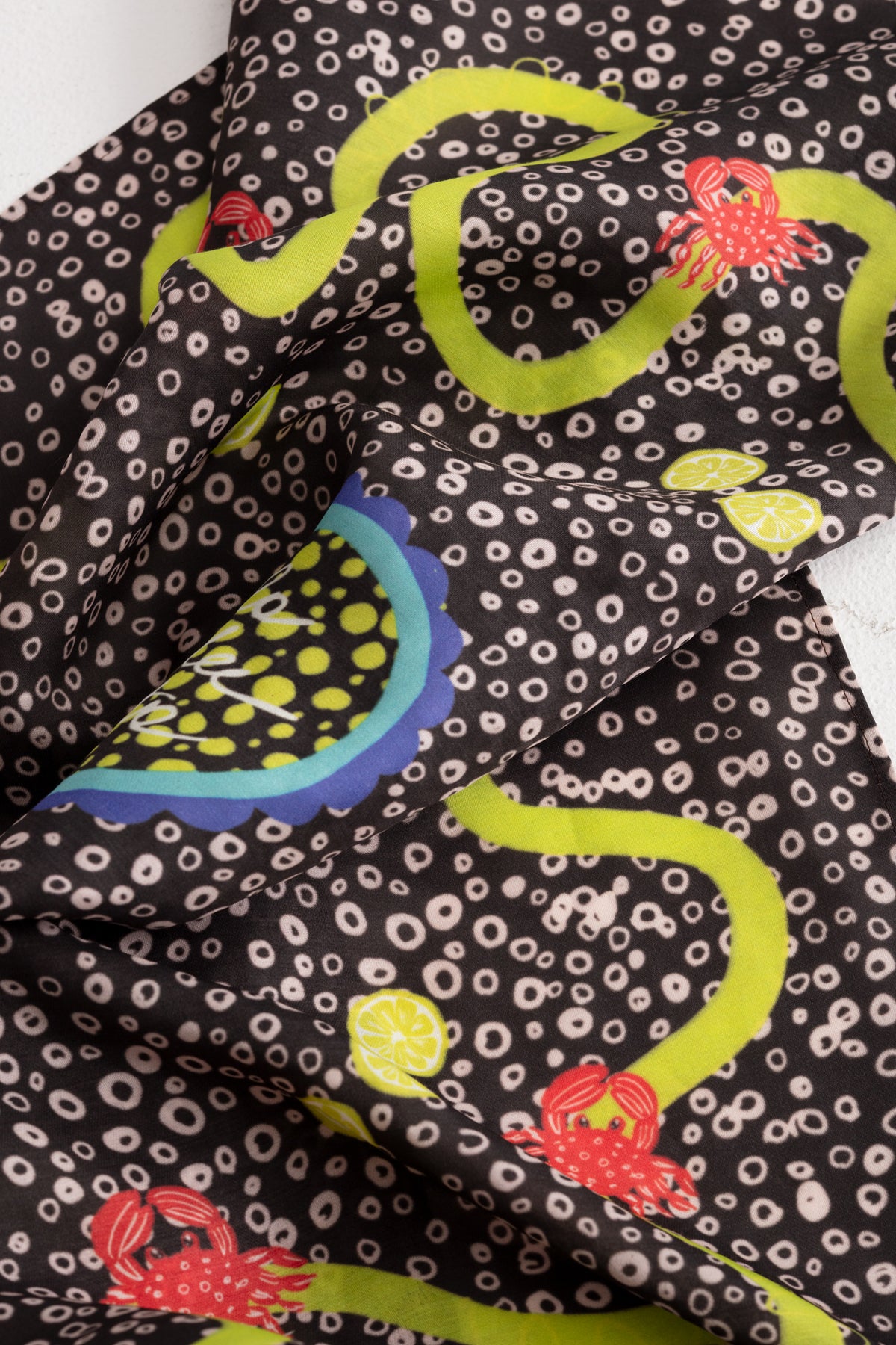 A close-up of a patterned silk and viscose fabric with a dark brown background, white circular spots, bright yellow-olive green wavy lines, red crabs, and blue and yellow circle motifs.