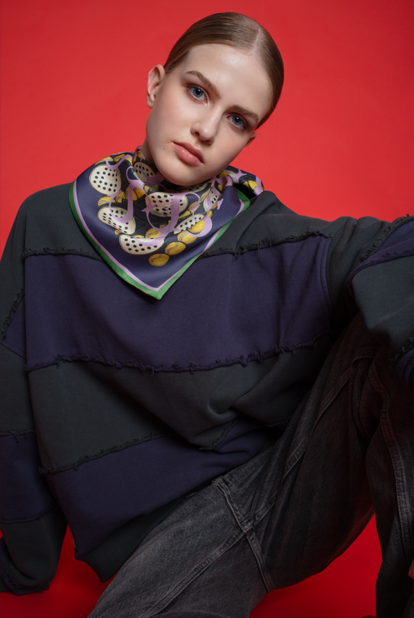 A studio fashion portrait of a person against a vibrant red background, wearing a MOVEMENT silk scarf tied neatly around the neck. The scarf features a complex geometric and organic pattern in shades of purple, yellow, and black, finished with a bright green border. The person is styled in a dark, oversized patchwork sweater and black jeans, leaning forward with a neutral expression, highlighting the scarf as a bold statement accessory in a modern, edgy outfit.