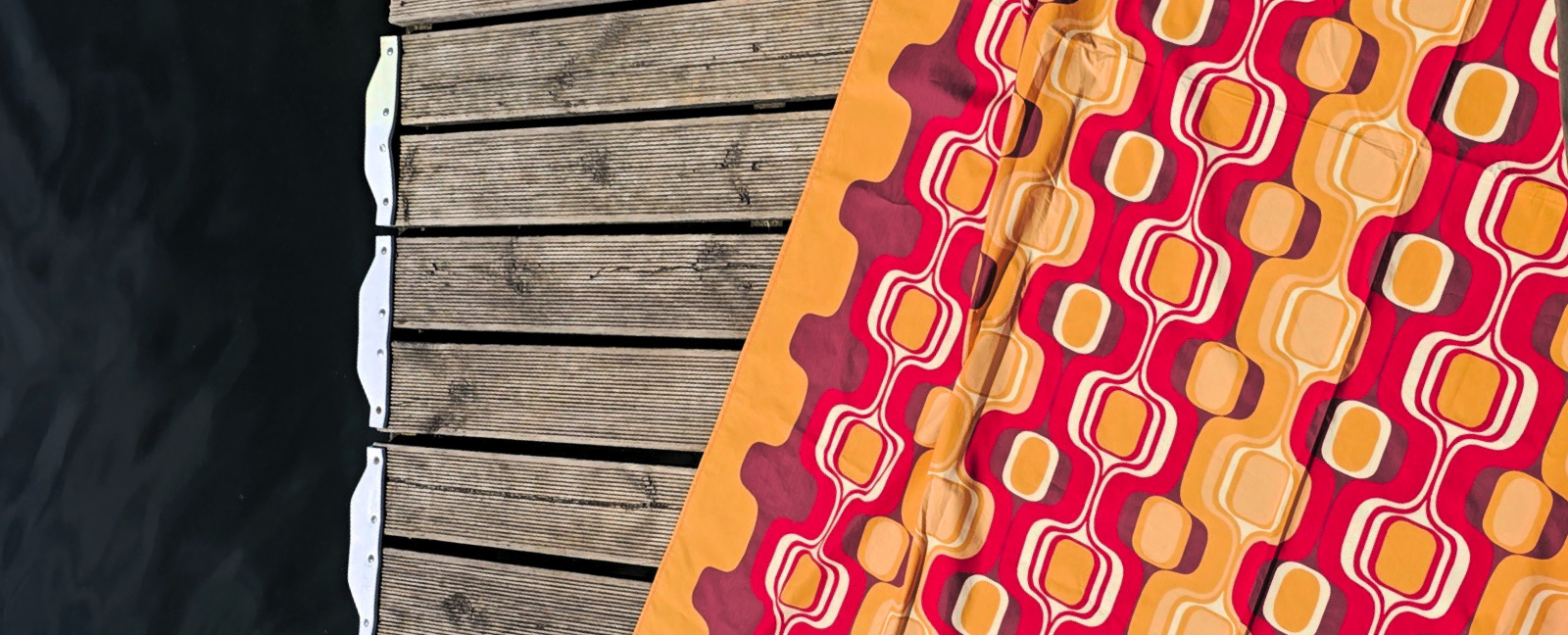 Bright orange and red retro-patterned cotton blanket spread on a wooden pier by the lake. Highlighting a vibrant summer vacation aesthetic and lightweight resort wear fashion accessory.