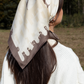 Outdoor lifestyle photography showing the back view of a brunette model wearing a cream silk headscarf with taupe and brown abstract geometric patterns, tied over long dark hair and styled with a chunky white knit sweater against a blurry forest background.