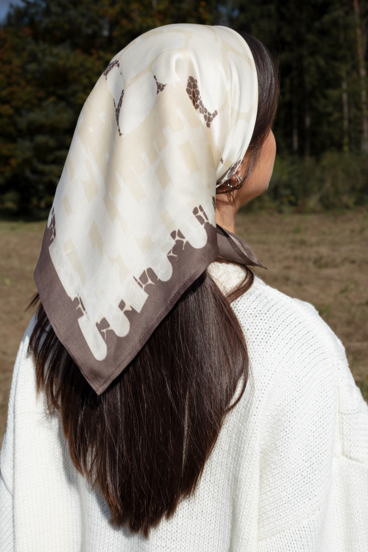 Outdoor lifestyle photography showing the back view of a brunette model wearing a cream silk headscarf with taupe and brown abstract geometric patterns, tied over long dark hair and styled with a chunky white knit sweater against a blurry forest background.