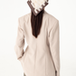 A model shown from the back wearing a light beige structured blazer, blue jeans, and a cream and brown patterned headscarf, set against a plain white studio background.