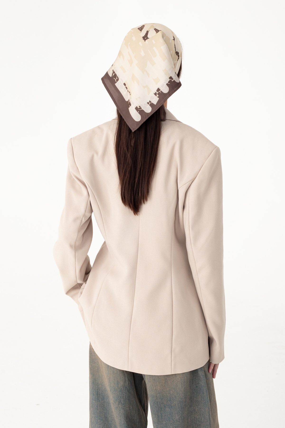 A model shown from the back wearing a light beige structured blazer, blue jeans, and a cream and brown patterned headscarf, set against a plain white studio background.