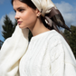 A woman outdoors wearing a cream cable-knit sweater and a cream satin silk scarf with brown patterned details, set against a blue sky and trees.
