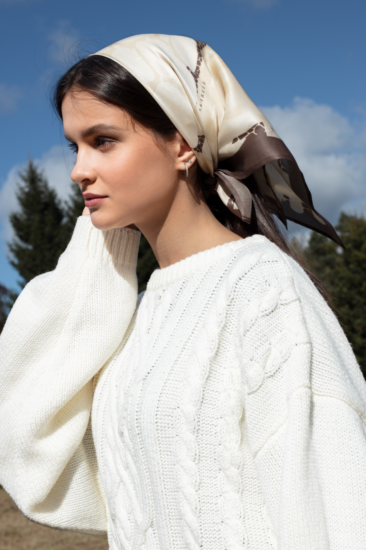 A woman outdoors wearing a cream cable-knit sweater and a cream satin silk scarf with brown patterned details, set against a blue sky and trees.