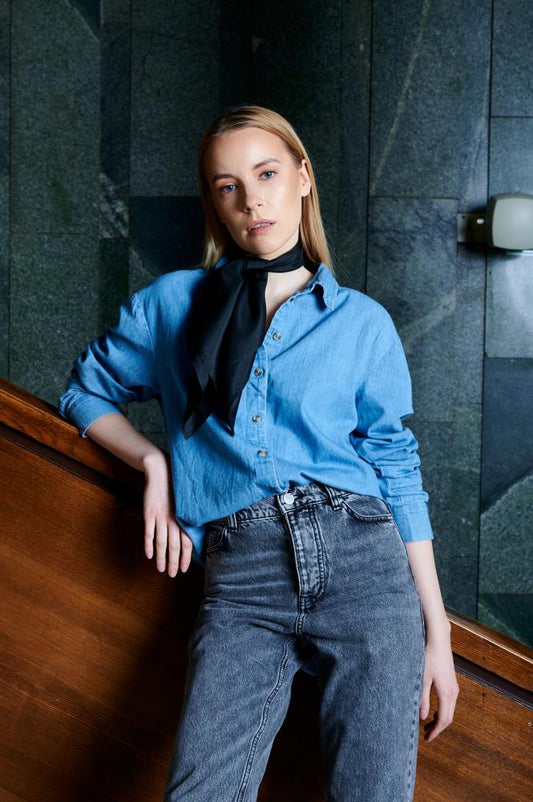 A model wearing a light blue denim shirt styled with a knotted silk neck scarf in a subtle greyish moss green shade, adding a sophisticated touch to a casual look.