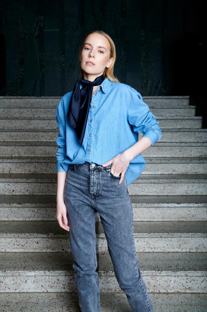 A model wearing a light blue denim shirt and grey jeans, styled with a knotted navy blue silk neck scarf for a classic, sophisticated everyday look.