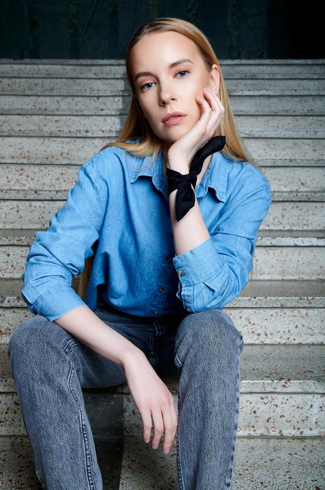 Young woman with long blonde hair, in a blue denim shirt and jeans, sitting on stairs. She rests her head on her hand that is styled with black silk pocket square/ handkerchief like a bracelet, looking contemplative.