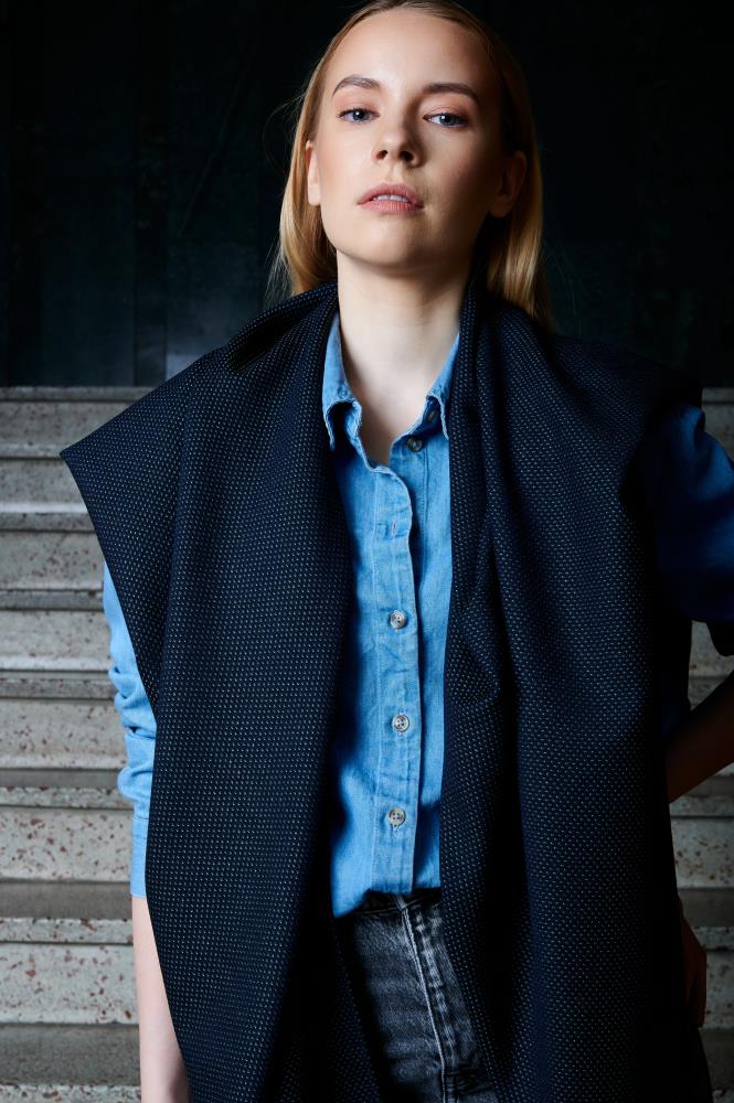 A woman poses standing on stairs, looking directly at the camera. She is wearing a light blue denim shirt and dark grey jeans, with a wide navy blue scarf featuring a fine textured pattern stylishly draped over her shoulders. The ends of the scarf hang freely in front, creating a layered and modern look. The staircase background and shadows add depth and an urban feel to the photo.