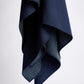 A vertical photo against a light background, featuring a dark blue scarf draped freely. The fabric has a fine, textured dotted pattern that appears lighter on the reverse side, creating a subtle contrast. The folds of the material highlight the product's lightness, softness, and high-quality stitching.