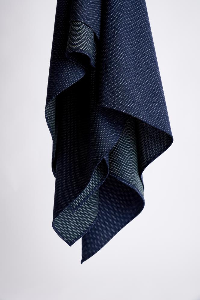 A vertical photo against a light background, featuring a dark blue scarf draped freely. The fabric has a fine, textured dotted pattern that appears lighter on the reverse side, creating a subtle contrast. The folds of the material highlight the product's lightness, softness, and high-quality stitching.