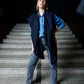A full-length photo of a woman posing on wide stone stairs in a dark interior. She is wearing a light blue denim shirt, grey jeans, and black pointed boots. A wide navy blue scarf is stylishly draped over her shoulders, falling freely in front to add a modern touch and volume to the outfit. The lighting is focused on the woman, highlighting the clothing details and creating a dramatic contrast with the dark background.