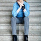 Young woman sits on concrete steps, wearing a blue denim shirt, gray jeans, and black boots. Her chin rests on her hands, one wrist is styled with silk pocket square/ handkerchief like a bracelet, conveying a calm, thoughtful expression.