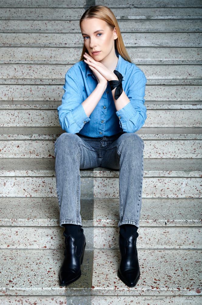 Young woman sits on concrete steps, wearing a blue denim shirt, gray jeans, and black boots. Her chin rests on her hands, one wrist is styled with silk pocket square/ handkerchief like a bracelet, conveying a calm, thoughtful expression.