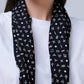 A close-up portrait of a woman from the lips to the chest, showcasing a dark scarf. The woman is wearing a simple white T-shirt, and draped around her neck is a black scarf decorated with small white zodiac sign symbols and stars. The ends of the scarf hang freely in front, creating a contrast with the light-colored garment and the neutral background.